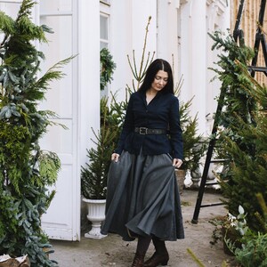 May include: A woman wearing a dark blue blazer with a black belt, a gray skirt, and brown boots. She is standing in front of a white building with greenery.