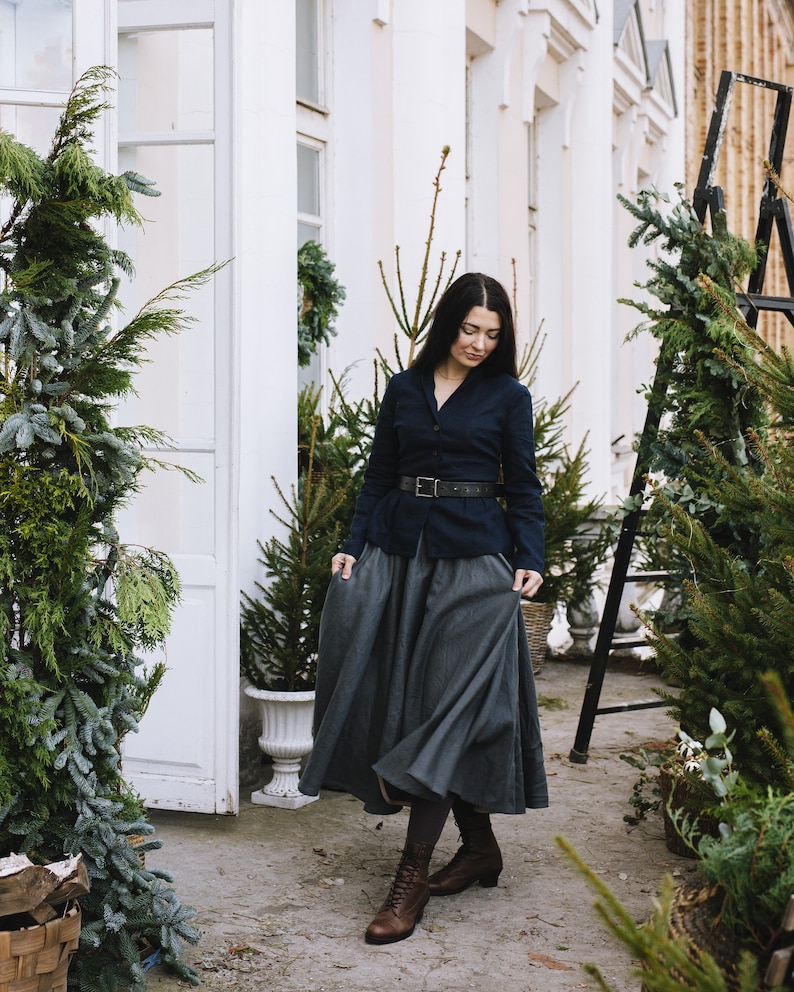 May include: A woman wearing a dark blue blazer with a black belt, a gray skirt, and brown boots. She is standing in front of a white building with greenery.
