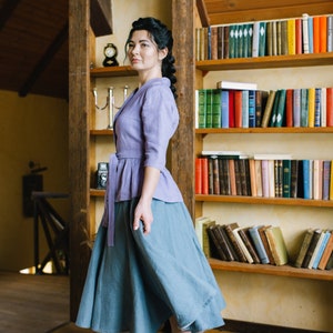 Op de afbeelding: Een vrouw draagt een lavendelkleurige linnen blouse met een geknoopte taille en een blauwe linnen rok. Ze staat in een kamer met een houten boekenkast vol boeken.