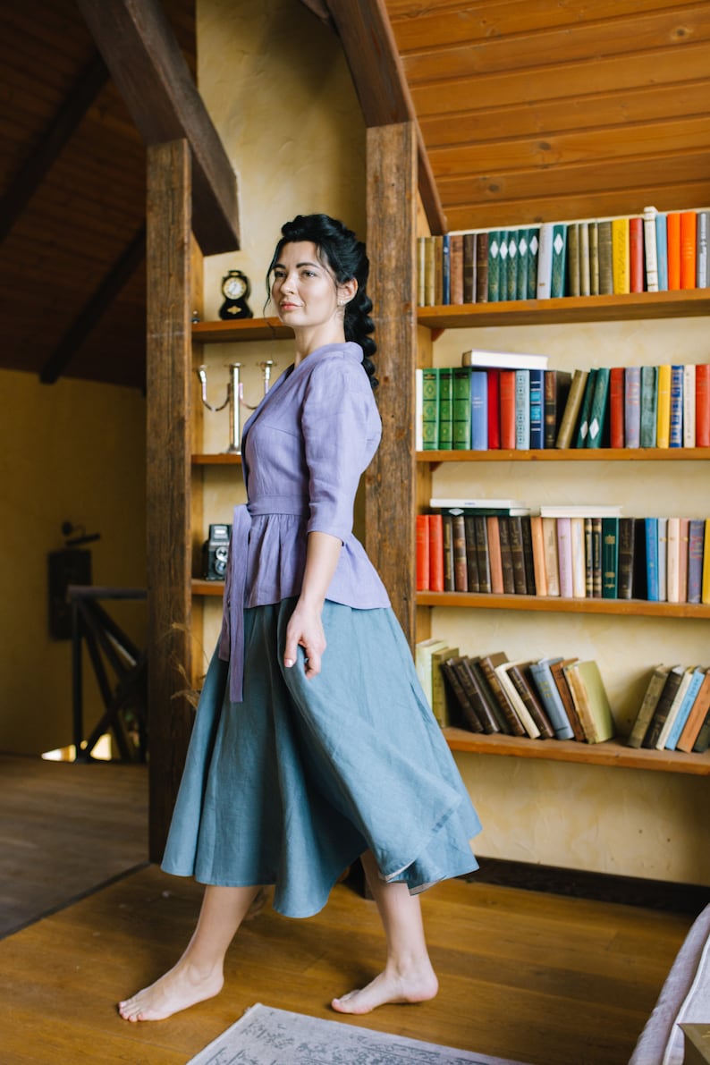 Op de afbeelding: Een vrouw draagt een lavendelkleurige linnen blouse met een geknoopte taille en een blauwe linnen rok. Ze staat in een kamer met een houten boekenkast vol boeken.