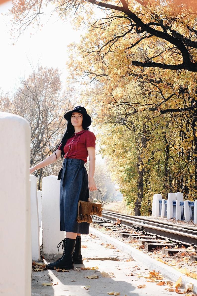 Puede incluir: Una persona con una camisa burdeos de manga corta, unos pantalones culotte vaqueros y un sombrero negro, de pie cerca de una valla blanca. Lleva un bolso marr&oacute;n con flecos y botas negras. El fondo muestra una v&iacute;a de tren y follaje oto&ntilde;al.
