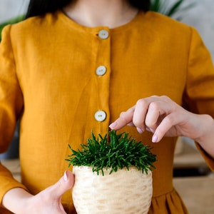 May include: A woman in a yellow linen dress holds a small potted succulent plant with green leaves. The pot is white with a textured surface.