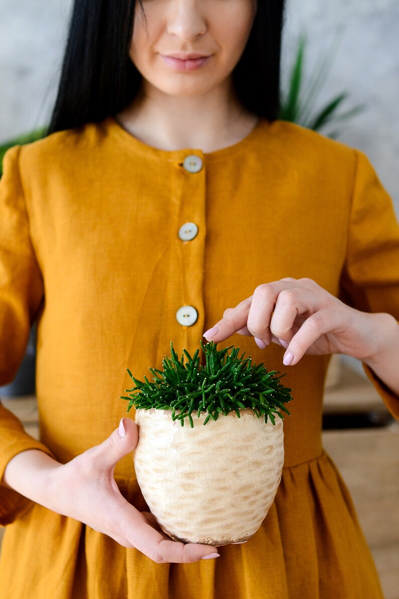 May include: A woman in a yellow linen dress holds a small potted succulent plant with green leaves. The pot is white with a textured surface.
