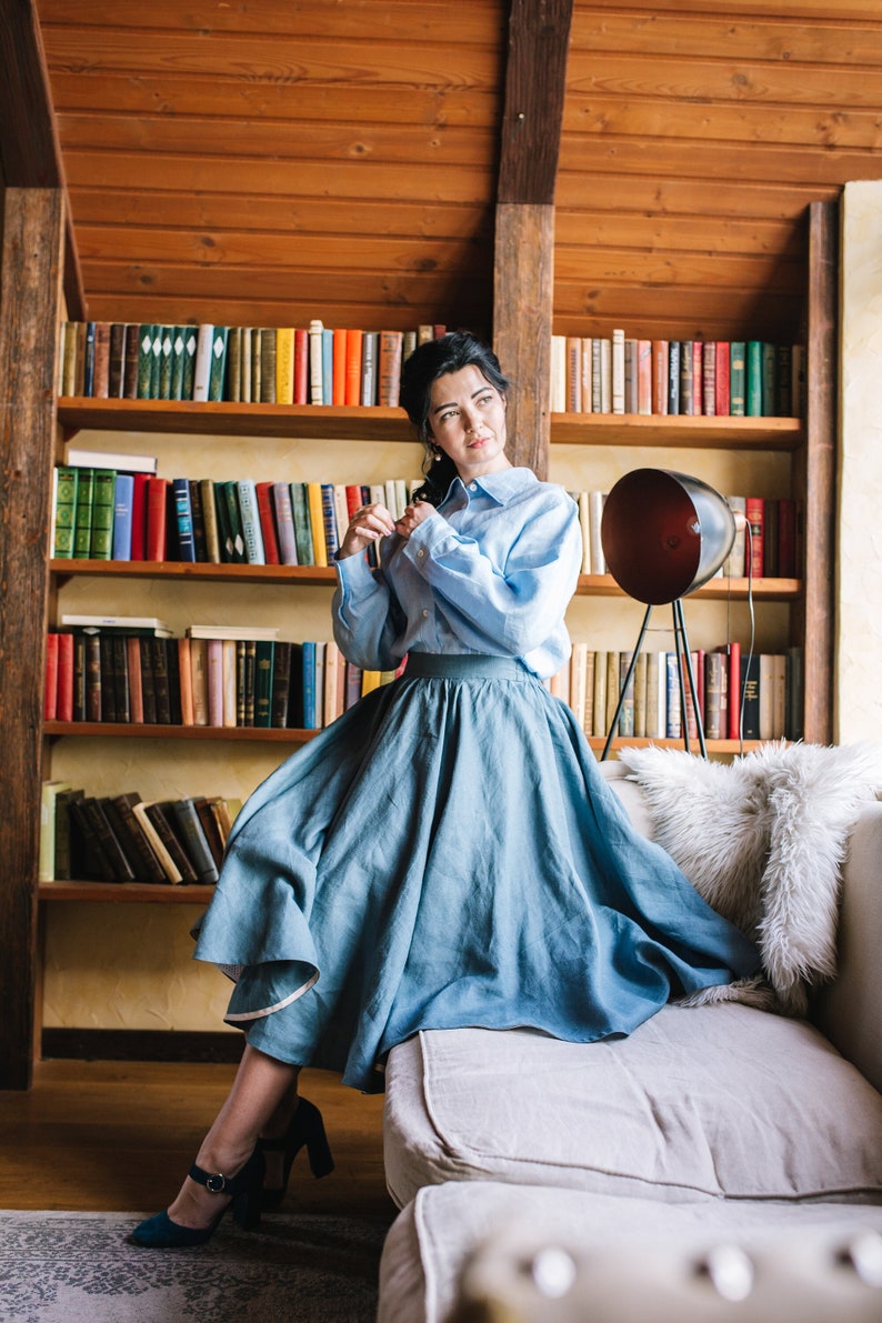 K&ouml;nnte beinhalten: Eine Frau in einem hellblauen Hemd und einem hellblauen Rock sitzt auf einem wei&szlig;en Sofa in einem Raum mit einem B&uuml;cherregal voller B&uuml;cher.