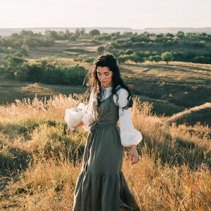 May include: A woman in a white shirt and a long, green, tiered dress walks through a field of tall grass. She is holding a bouquet of white flowers.