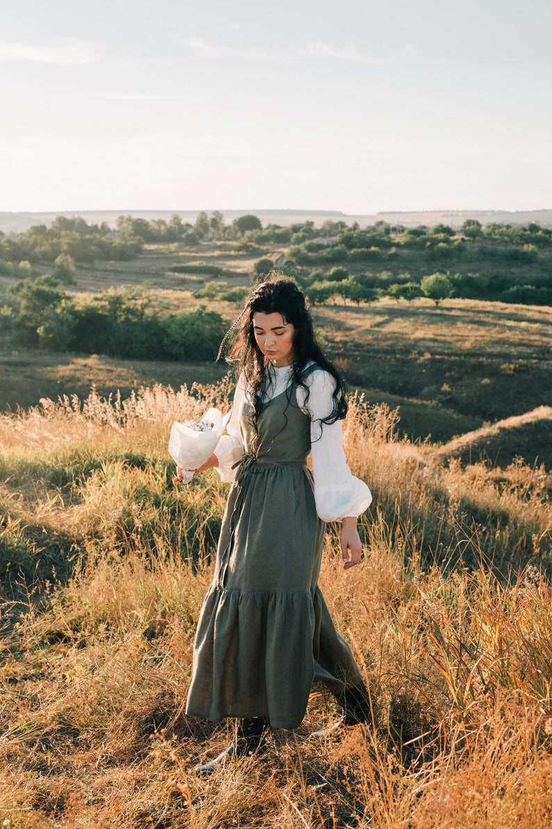 May include: A woman in a white shirt and a long, green, tiered dress walks through a field of tall grass. She is holding a bouquet of white flowers.