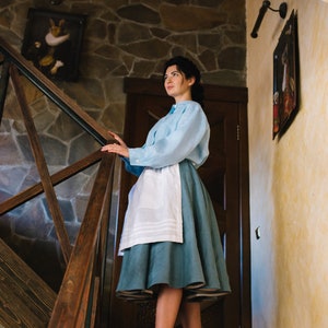 K&ouml;nnte beinhalten: Eine Frau in einem hellblauen Hemd und einem blauen Rock mit einer wei&szlig;en Sch&uuml;rze steht auf einer Holztreppe. Sie tr&auml;gt gr&uuml;ne Schuhe mit Absatz und schaut nach oben.