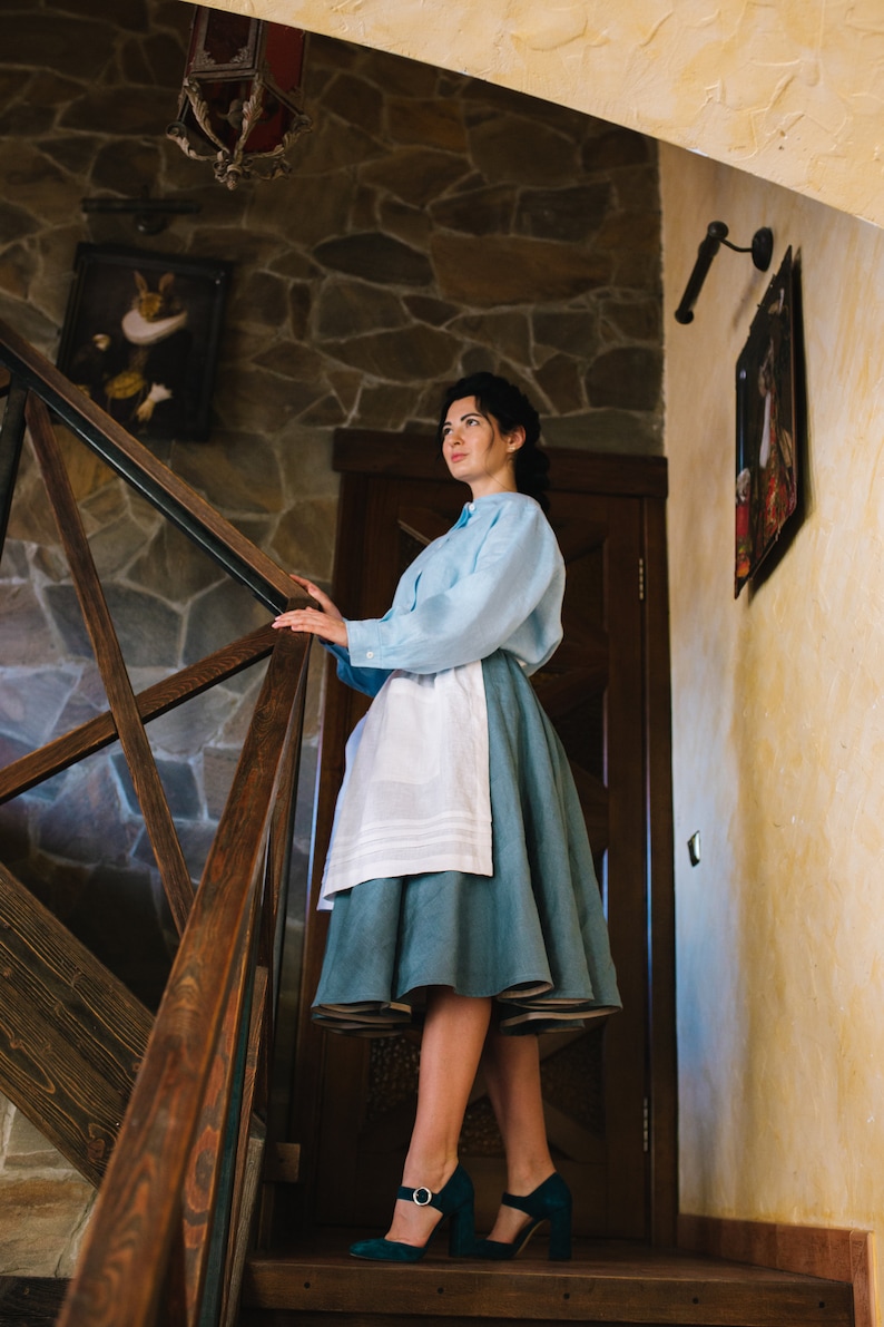 K&ouml;nnte beinhalten: Eine Frau in einem hellblauen Hemd und einem blauen Rock mit einer wei&szlig;en Sch&uuml;rze steht auf einer Holztreppe. Sie tr&auml;gt gr&uuml;ne Schuhe mit Absatz und schaut nach oben.