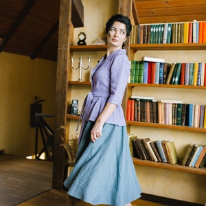 Op de afbeelding: Een vrouw in een lavendelkleurig blouse met een strik in de taille en een blauwe rok staat in een kamer met een houten boekenkast vol boeken.