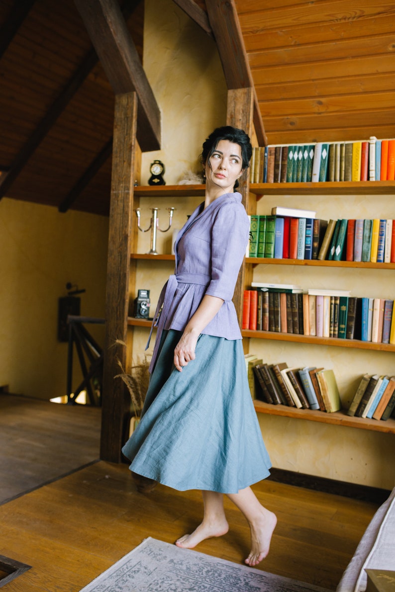 Op de afbeelding: Een vrouw in een lavendelkleurig blouse met een strik in de taille en een blauwe rok staat in een kamer met een houten boekenkast vol boeken.