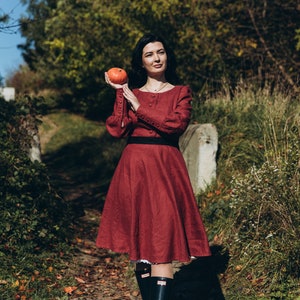 May include: A woman in a maroon dress with a black belt and black boots holds a small orange pumpkin in her hand. She is standing on a path in a forest.