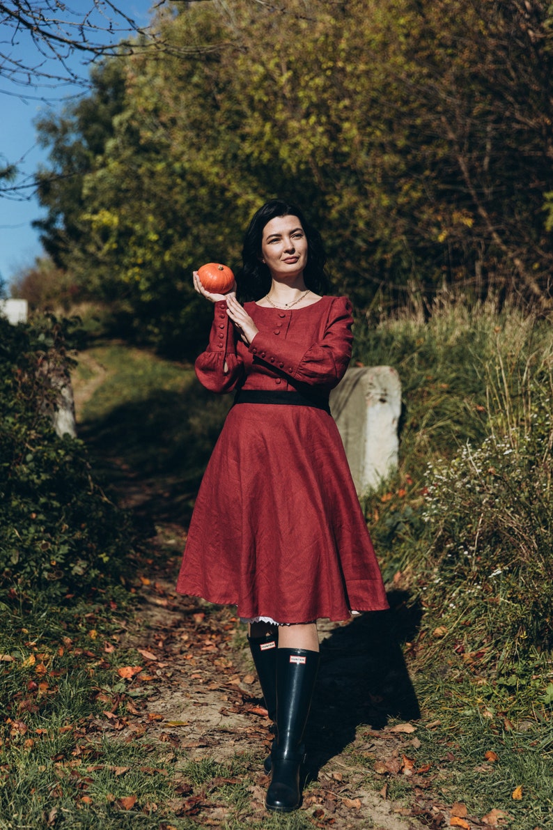 May include: A woman in a maroon dress with a black belt and black boots holds a small orange pumpkin in her hand. She is standing on a path in a forest.