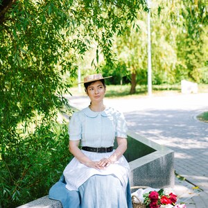 K&ouml;nnte beinhalten: Eine Frau in einem hellblauen Kleid mit wei&szlig;em Kragen und einem Strohhut sitzt auf einer Steinbank. Sie h&auml;lt einen Strau&szlig; roter und wei&szlig;er Blumen in einem Weidenkorb.