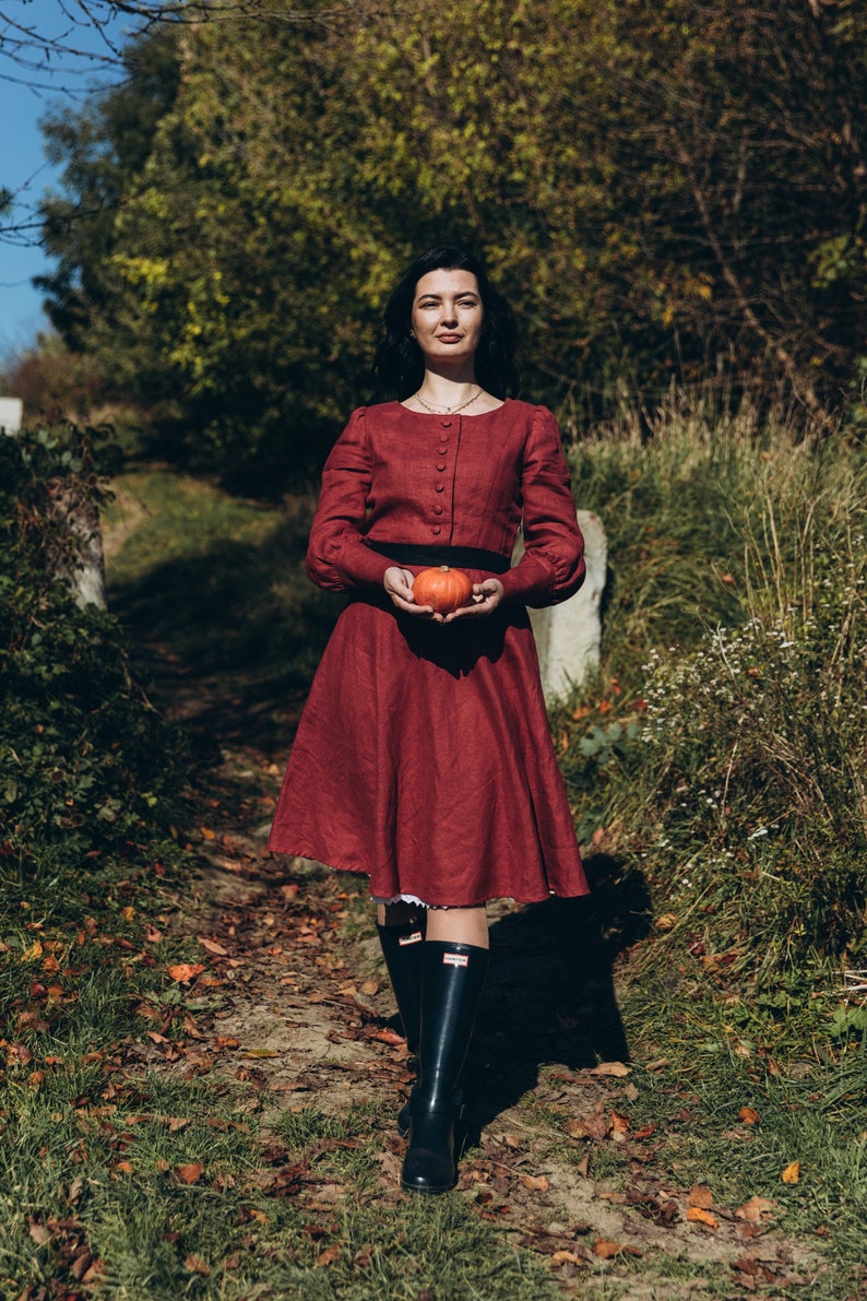 May include: A woman in a burgundy dress holds a small orange pumpkin in a forest setting. She is wearing black rain boots.