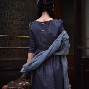 Puede incluir: Una mujer con un vestido azul y un gorro gris se encuentra frente a una puerta, sosteniendo una maleta marr&oacute;n. Lleva una bufanda azul drapeada sobre su hombro.