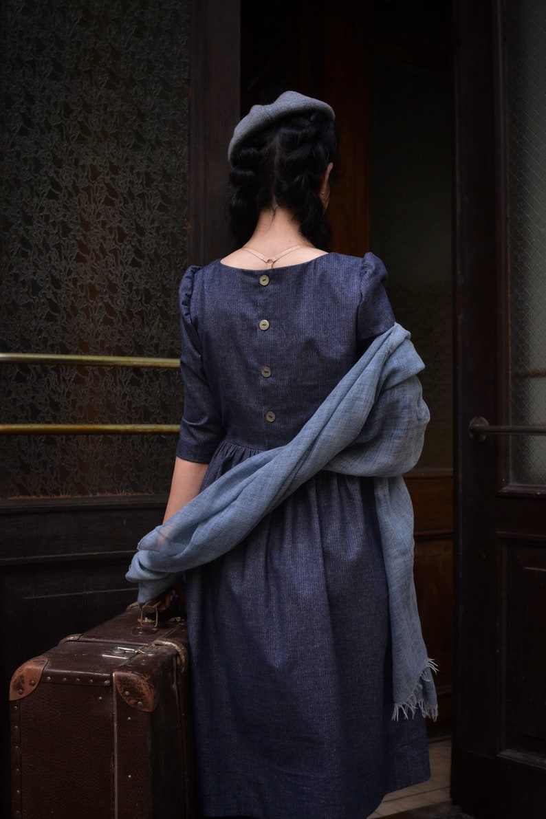 Puede incluir: Una mujer con un vestido azul y un gorro gris se encuentra frente a una puerta, sosteniendo una maleta marr&oacute;n. Lleva una bufanda azul drapeada sobre su hombro.