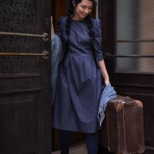 Puede incluir: Una mujer con un vestido de mezclilla azul y una boina gris se encuentra en una puerta sosteniendo una maleta vintage marr&oacute;n. Lleva medias negras y botas negras.