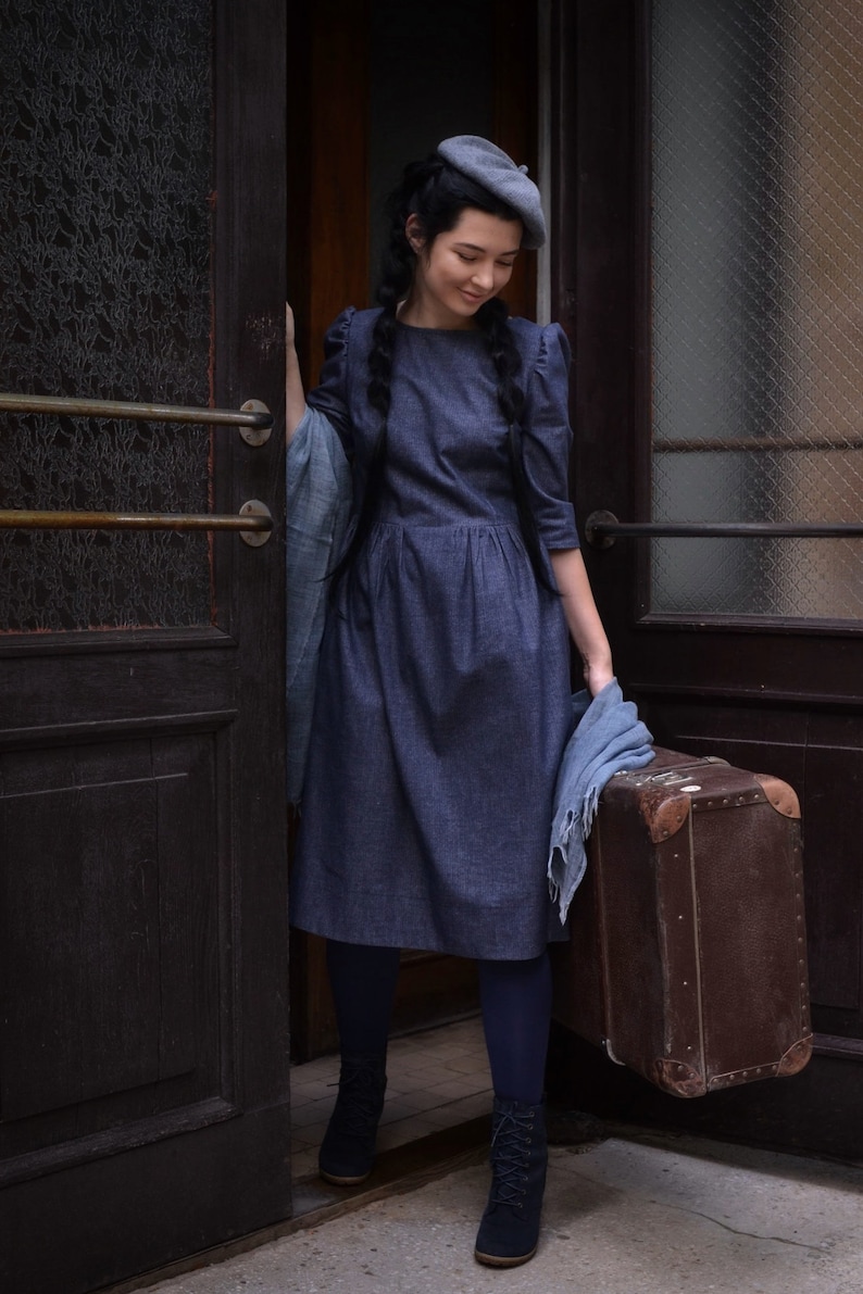 Puede incluir: Una mujer con un vestido de mezclilla azul y una boina gris se encuentra en una puerta sosteniendo una maleta vintage marr&oacute;n. Lleva medias negras y botas negras.