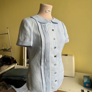 Puede incluir: Blusa celeste de manga corta con cuello redondo y pliegues verticales en la parte delantera. La blusa tiene una fila de botones oscuros y se muestra en un maniqu&iacute;. El fondo es una pared amarillo claro y una mesa blanca.