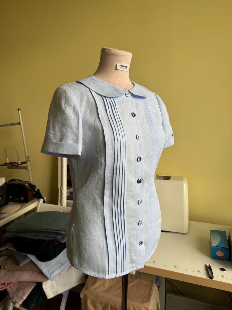 Puede incluir: Blusa celeste de manga corta con cuello redondo y pliegues verticales en la parte delantera. La blusa tiene una fila de botones oscuros y se muestra en un maniqu&iacute;. El fondo es una pared amarillo claro y una mesa blanca.