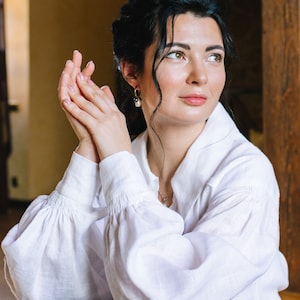May include: A woman in a white linen shirt with gathered sleeves and a blue skirt. The shirt has a classic collar and buttoned cuffs. She is wearing pearl earrings and a silver necklace.