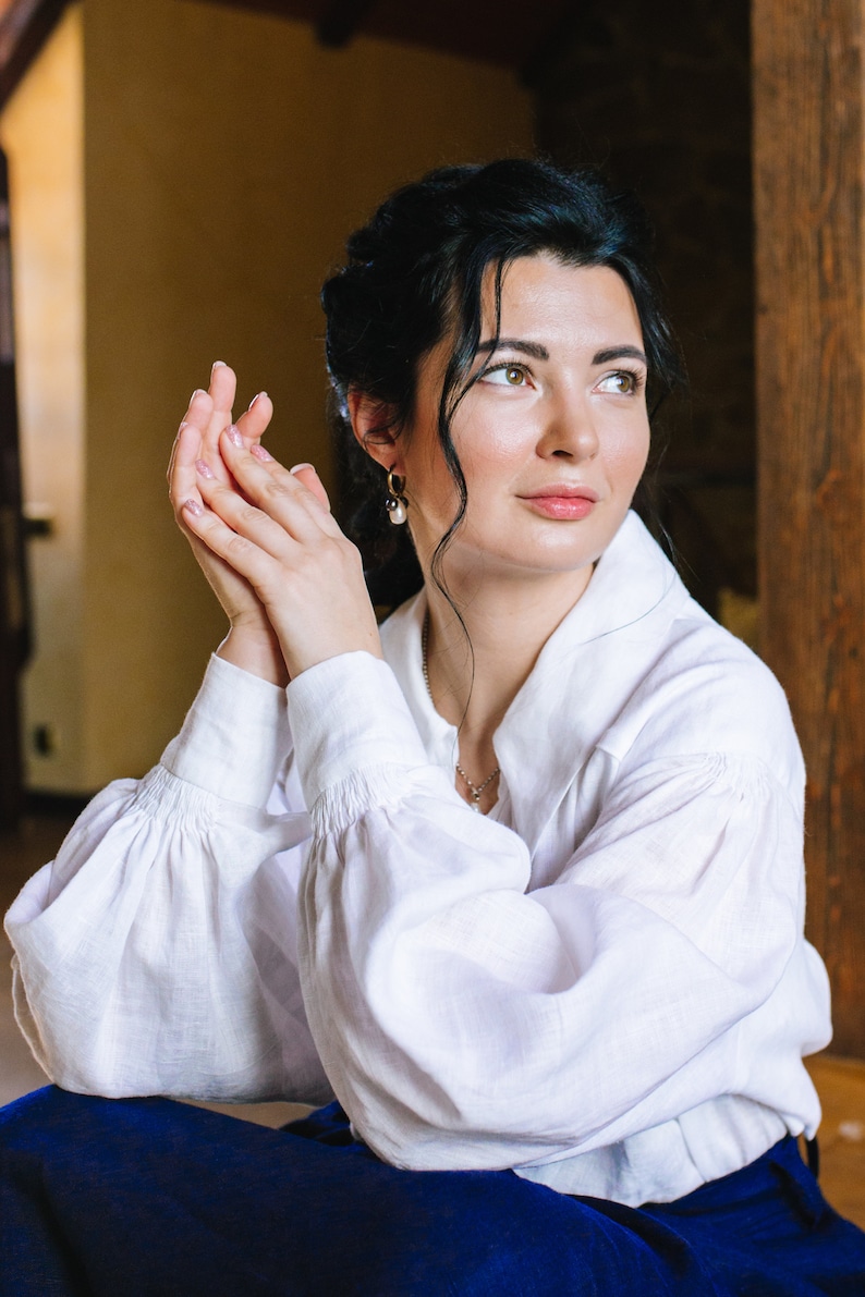 May include: A woman in a white linen shirt with gathered sleeves and a blue skirt. The shirt has a classic collar and buttoned cuffs. She is wearing pearl earrings and a silver necklace.