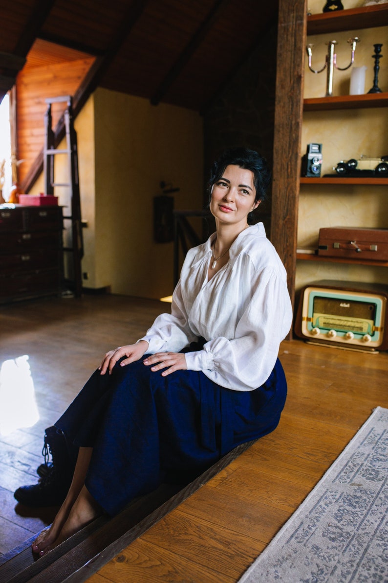 May include: A woman in a white blouse and a navy blue skirt sits on wooden steps. The room has a vintage feel, with a wooden bookshelf, a radio, and a vintage suitcase.