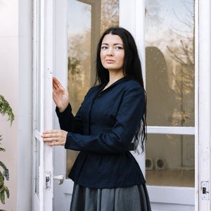 May include: A woman wearing a dark blue fitted jacket with a grey skirt, standing in front of a white door with glass panels.