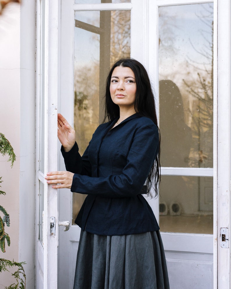 May include: A woman wearing a dark blue fitted jacket with a grey skirt, standing in front of a white door with glass panels.