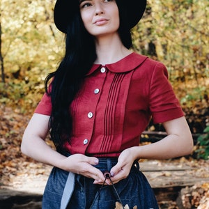 Puede incluir: Una persona con sombrero negro y camisa roja con cuello y botones. Sostiene una cesta de alambre llena de calabazas y hojas de oto&ntilde;o. El fondo muestra un bosque con follaje amarillo y marr&oacute;n.