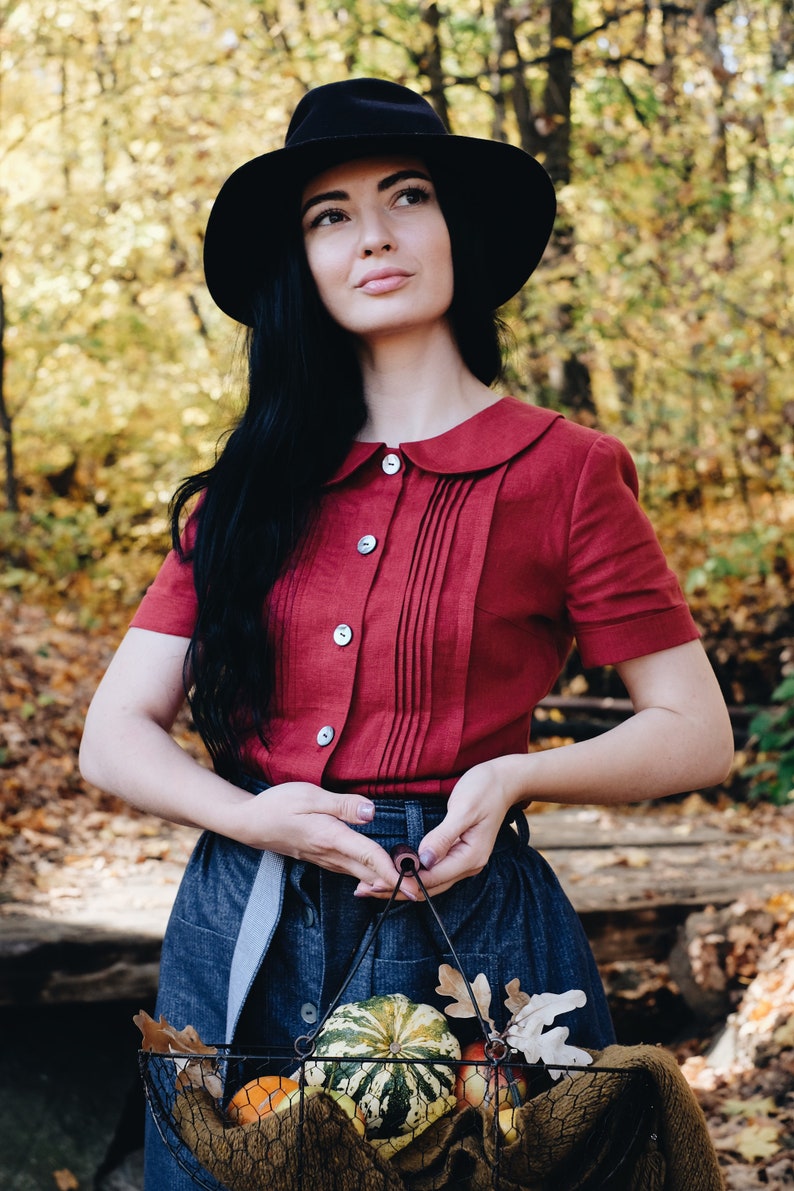 Puede incluir: Una persona con sombrero negro y camisa roja con cuello y botones. Sostiene una cesta de alambre llena de calabazas y hojas de oto&ntilde;o. El fondo muestra un bosque con follaje amarillo y marr&oacute;n.