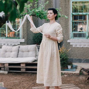 Pu&ograve; includere: Una donna con un abito di lino color crema si trova su una passerella di legno in un giardino. Sta tenendo un nastro di stoffa bianco e guardando di lato.