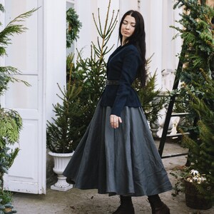 May include: A woman wearing a dark blue blazer with a grey skirt and brown boots. She is standing in front of a white building with greenery.