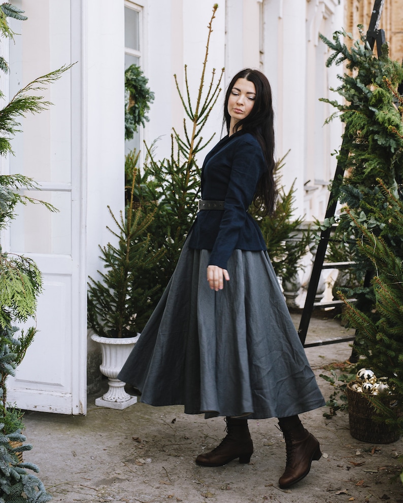 May include: A woman wearing a dark blue blazer with a grey skirt and brown boots. She is standing in front of a white building with greenery.