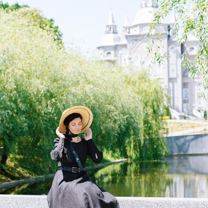 May include: A woman wearing a black dress and a straw hat sits on a stone wall with a wicker basket and a bouquet of pink and white flowers. She is wearing brown boots and has her eyes closed.