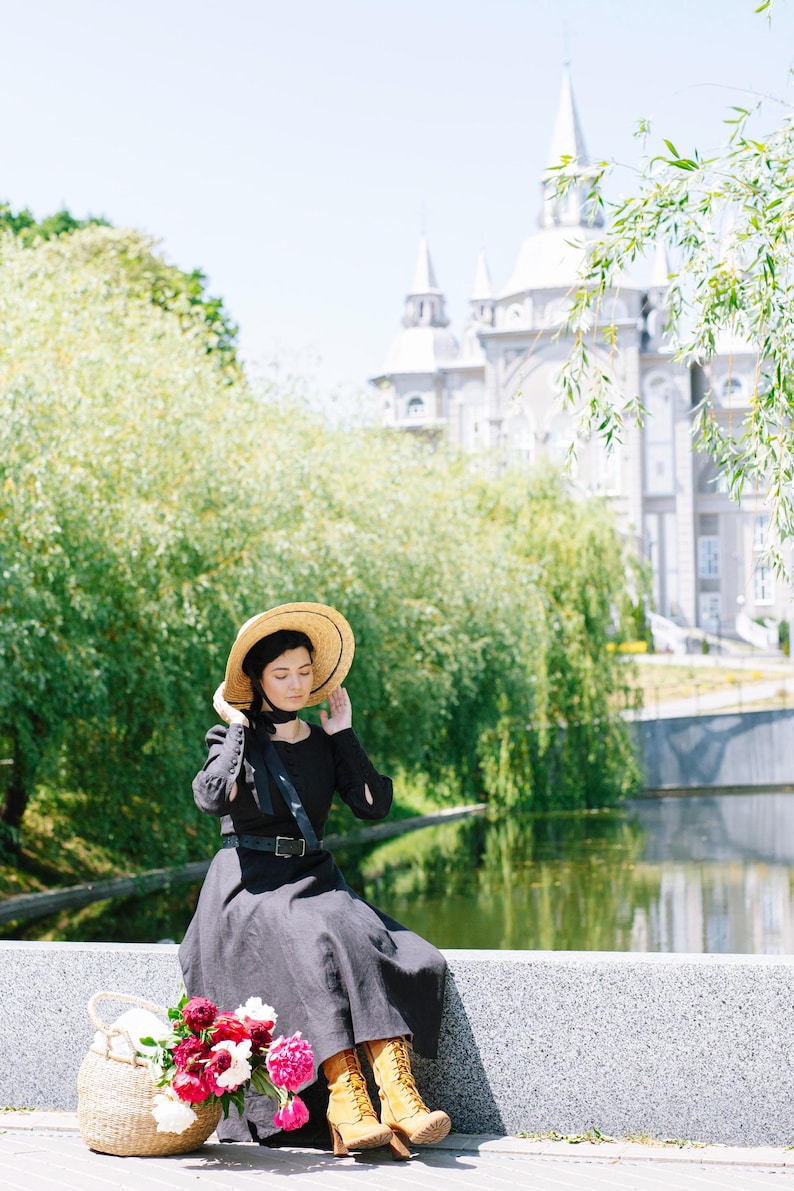 May include: A woman wearing a black dress and a straw hat sits on a stone wall with a wicker basket and a bouquet of pink and white flowers. She is wearing brown boots and has her eyes closed.