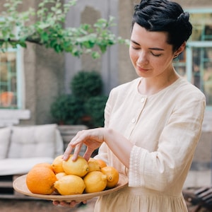 Pu&ograve; includere: Una donna con un vestito beige chiaro tiene una ciotola di legno piena di limoni e arance. La ciotola &egrave; rotonda e ha una finitura in legno naturale. I limoni sono gialli e le arance sono arancioni.
