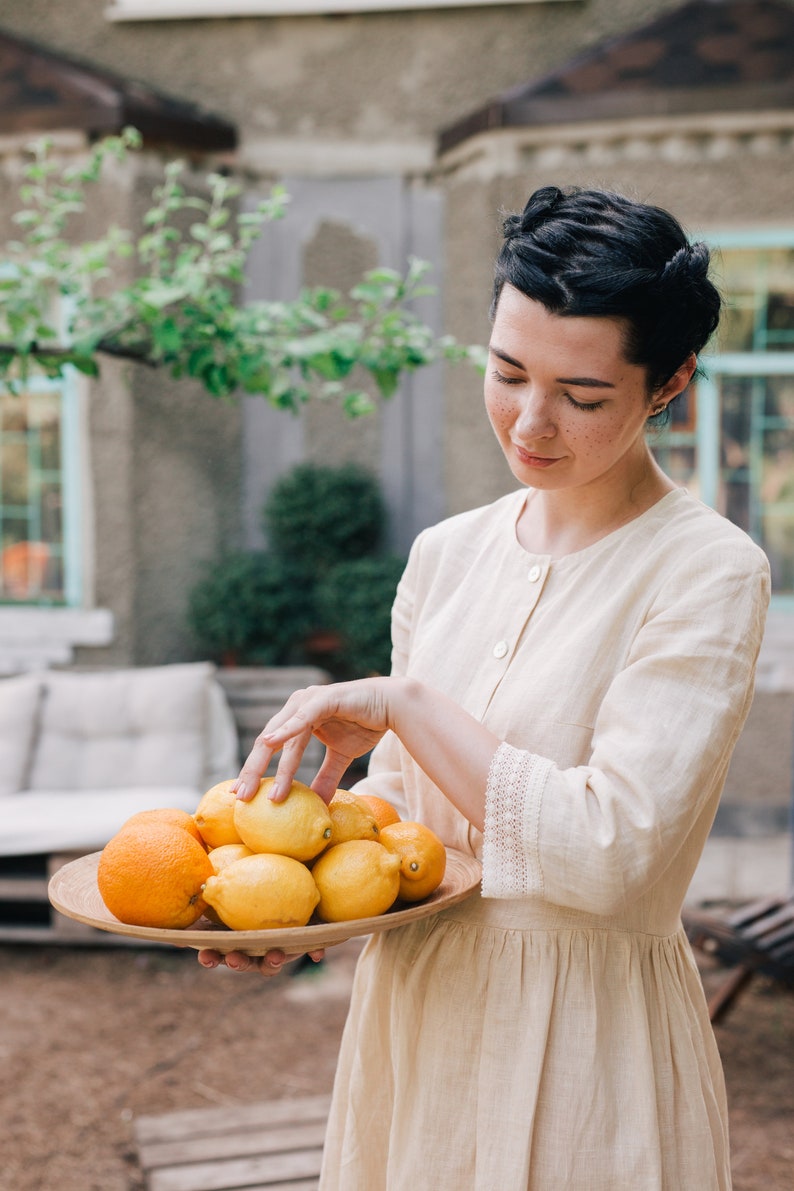 Pu&ograve; includere: Una donna con un vestito beige chiaro tiene una ciotola di legno piena di limoni e arance. La ciotola &egrave; rotonda e ha una finitura in legno naturale. I limoni sono gialli e le arance sono arancioni.