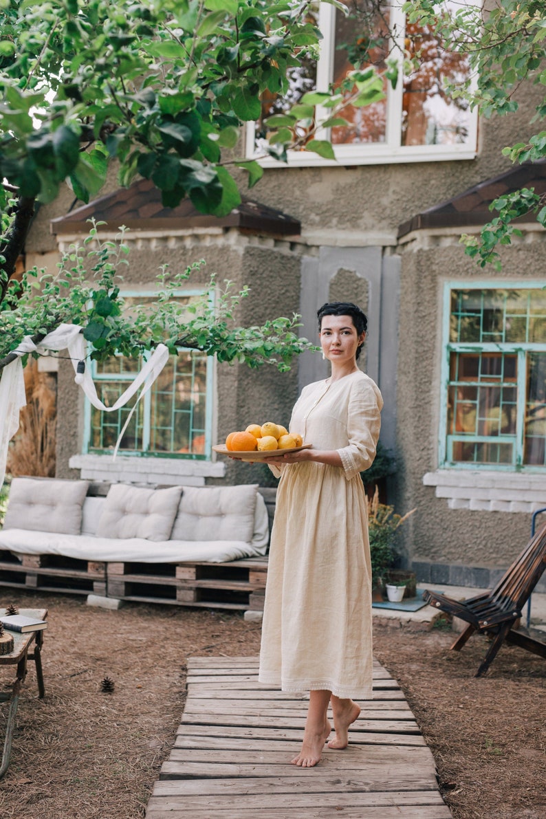 Pu&ograve; includere: Una donna con un vestito di lino bianco tiene un vassoio di legno con limoni e arance freschi. &Egrave; in piedi su una passerella di legno in un giardino.