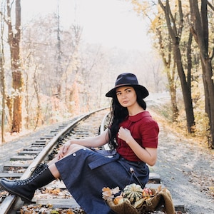Puede incluir: Una persona con sombrero oscuro, top rojo y falda vaquera sentada en v&iacute;as de tren. Una cesta con calabazas y una bufanda descansa cerca. La escena se desarrolla en un bosque con follaje oto&ntilde;al.
