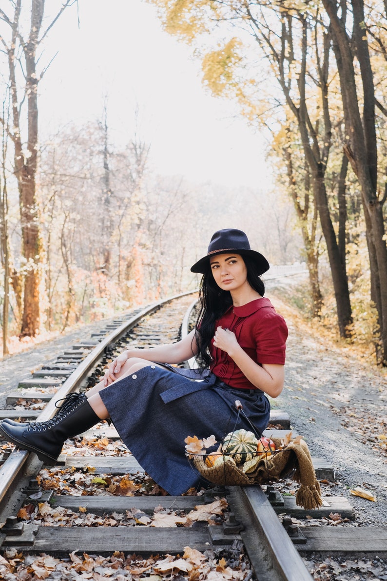 Puede incluir: Una persona con sombrero oscuro, top rojo y falda vaquera sentada en v&iacute;as de tren. Una cesta con calabazas y una bufanda descansa cerca. La escena se desarrolla en un bosque con follaje oto&ntilde;al.