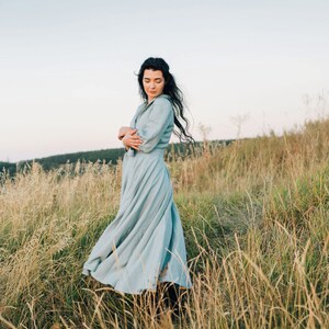 Puede incluir: Una mujer con un vestido azul claro se encuentra de pie en un campo de hierba alta. El vestido tiene una falda fluida y mangas largas. La mujer est&aacute; mirando hacia el suelo.