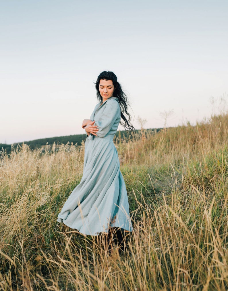 Puede incluir: Una mujer con un vestido azul claro se encuentra de pie en un campo de hierba alta. El vestido tiene una falda fluida y mangas largas. La mujer est&aacute; mirando hacia el suelo.