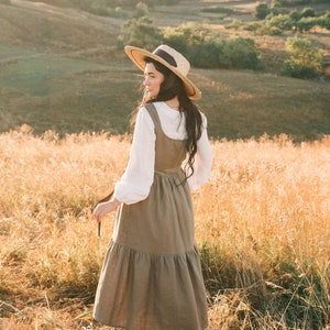 May include: A woman wearing a straw hat and a brown linen dress stands in a field of tall grass. The sun is shining and the sky is blue.