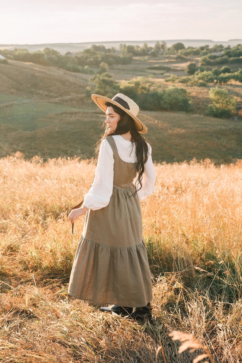 May include: A woman wearing a straw hat and a brown linen dress stands in a field of tall grass. The sun is shining and the sky is blue.