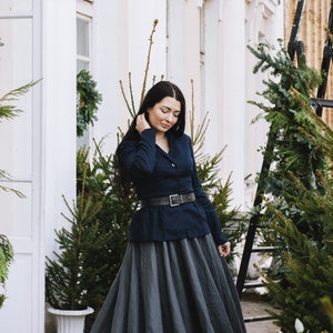 May include: A woman wearing a dark blue blazer with a black belt and a long, gray skirt. She is standing in front of a white building with green plants.