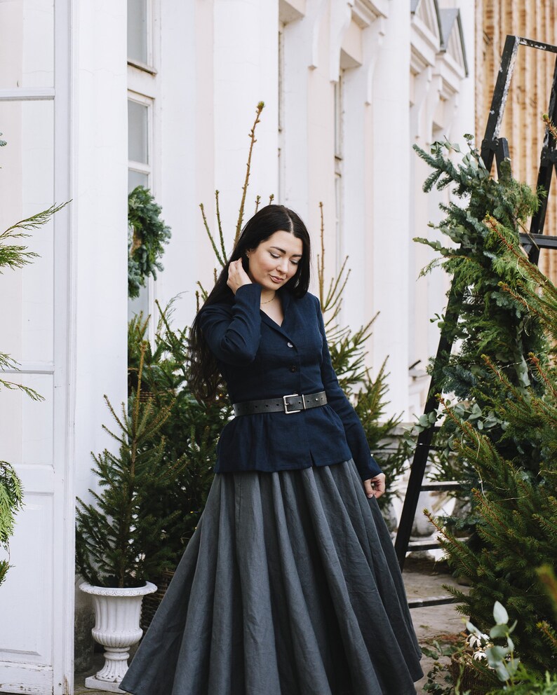 May include: A woman wearing a dark blue blazer with a black belt and a long, gray skirt. She is standing in front of a white building with green plants.