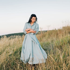 Puede incluir: Una mujer con un vestido de lino azul claro se encuentra en un campo de hierba alta. El vestido tiene una falda amplia y un cierre de botones en la parte delantera.