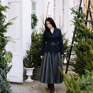 May include: A woman wearing a dark blue blazer with a black belt and a gray skirt, standing in front of a white building with potted evergreen trees.
