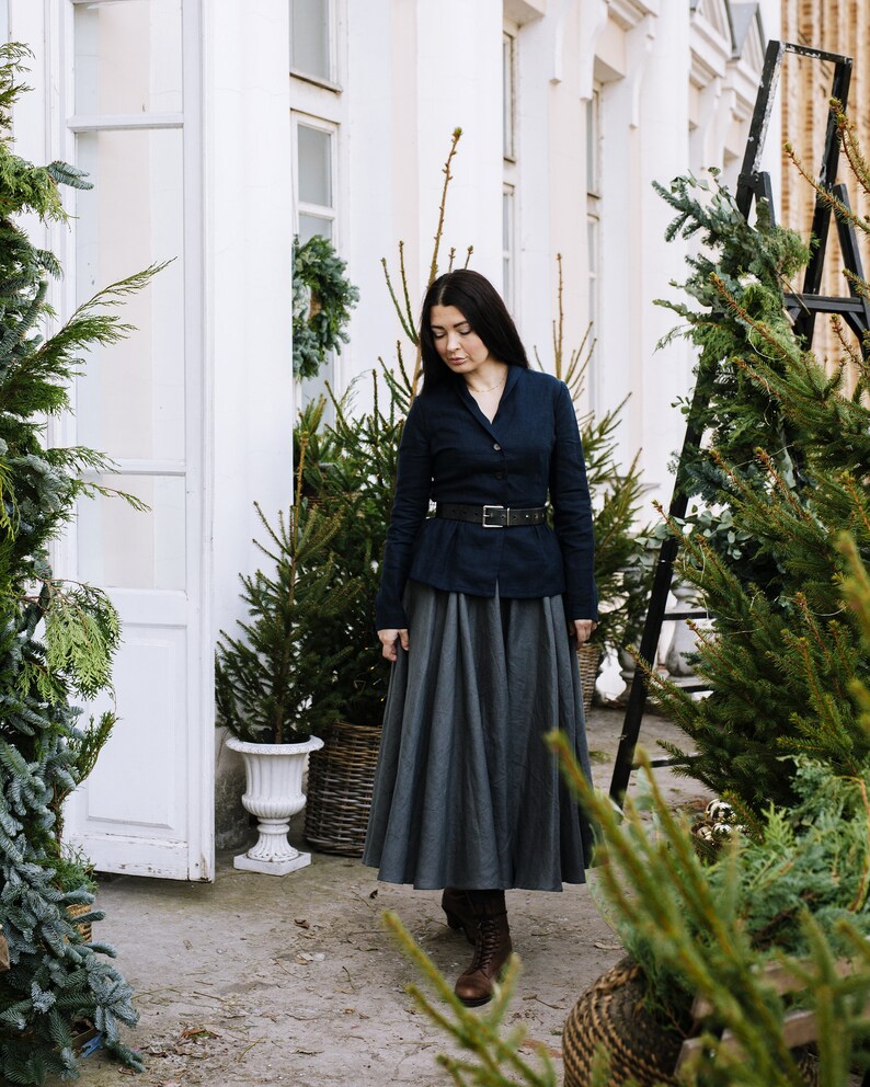 May include: A woman wearing a dark blue blazer with a black belt and a gray skirt, standing in front of a white building with potted evergreen trees.